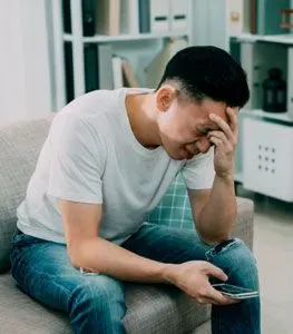 Image of a young man sitting on the couch holding a smart phone with his head in his hand looking exhausted